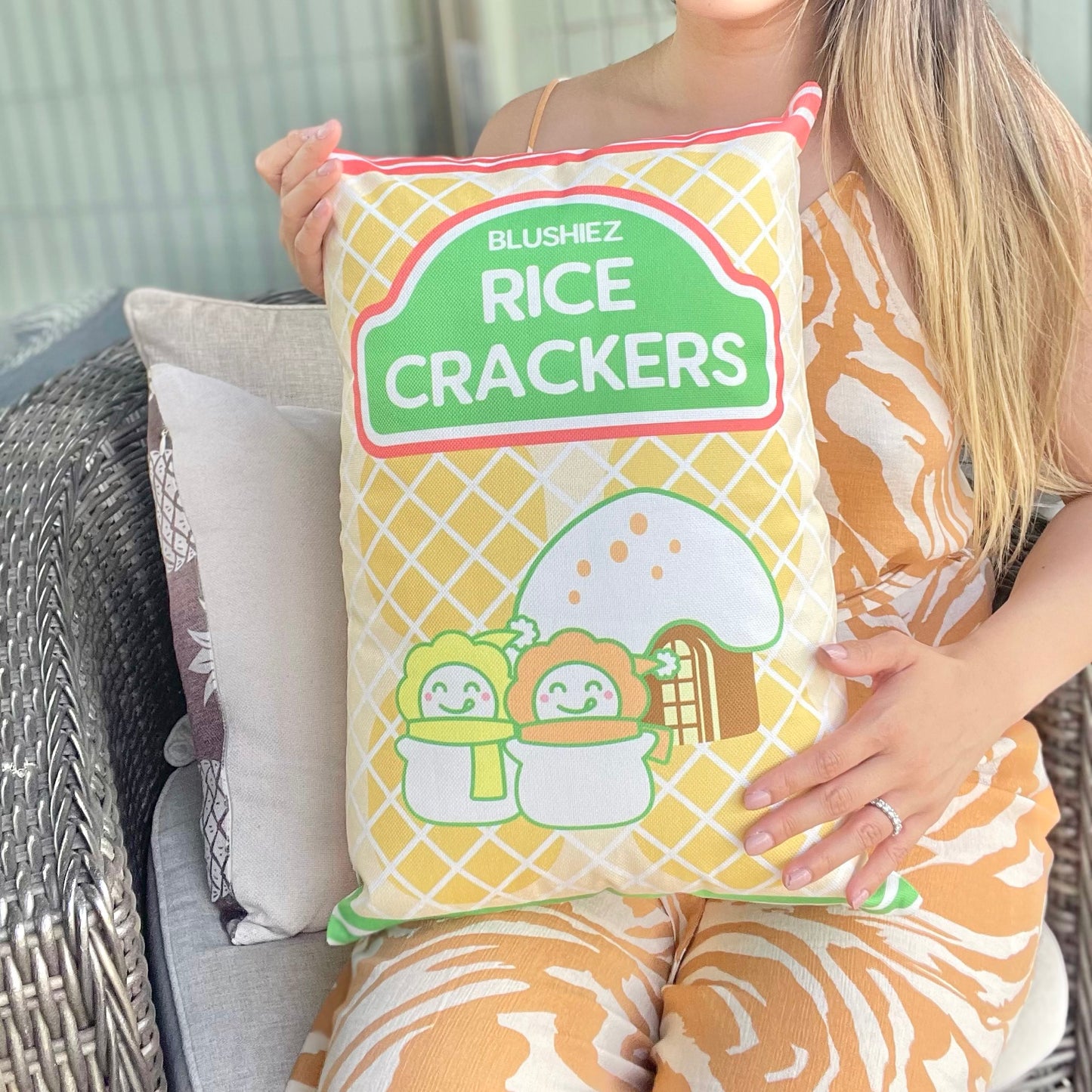 Pillow with a festive snack-inspired design, held by a woman in her lap. The pillow features a light orange background resembling rice crackers, with orange stripes at the top and green stripes at the bottom. The design includes a charming scene of two snowmen in front of a house with a snowy roof, evoking a festive and wintery atmosphere.