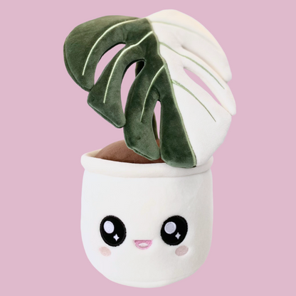 Plush toy of a potted plant with a white pot and a cute open-mouthed smiling face on the pot. The plant is a single leaf of a Monstera deliciosa albo variegata, featuring a split color pattern. The left half of the leaf is green, while the right half is white.