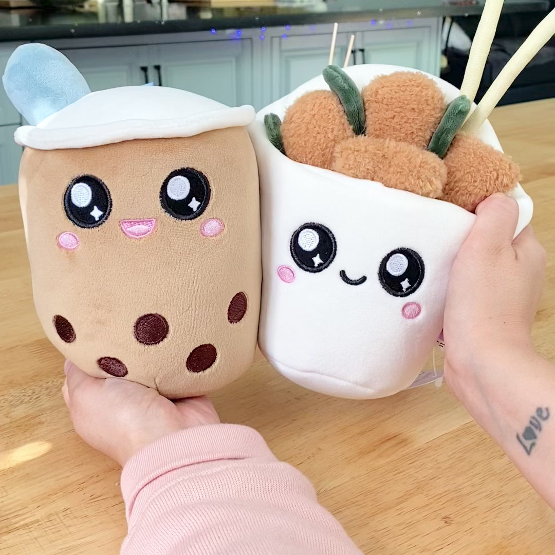 A video showcasing plush toys of a boba drink and popcorn chicken characters with magnetic features. The video demonstrates the toys being pulled apart and brought back together, showcasing the magnetic aspect of the toys. In the background, there is a cup of actual boba milk tea and a box of popcorn chicken.