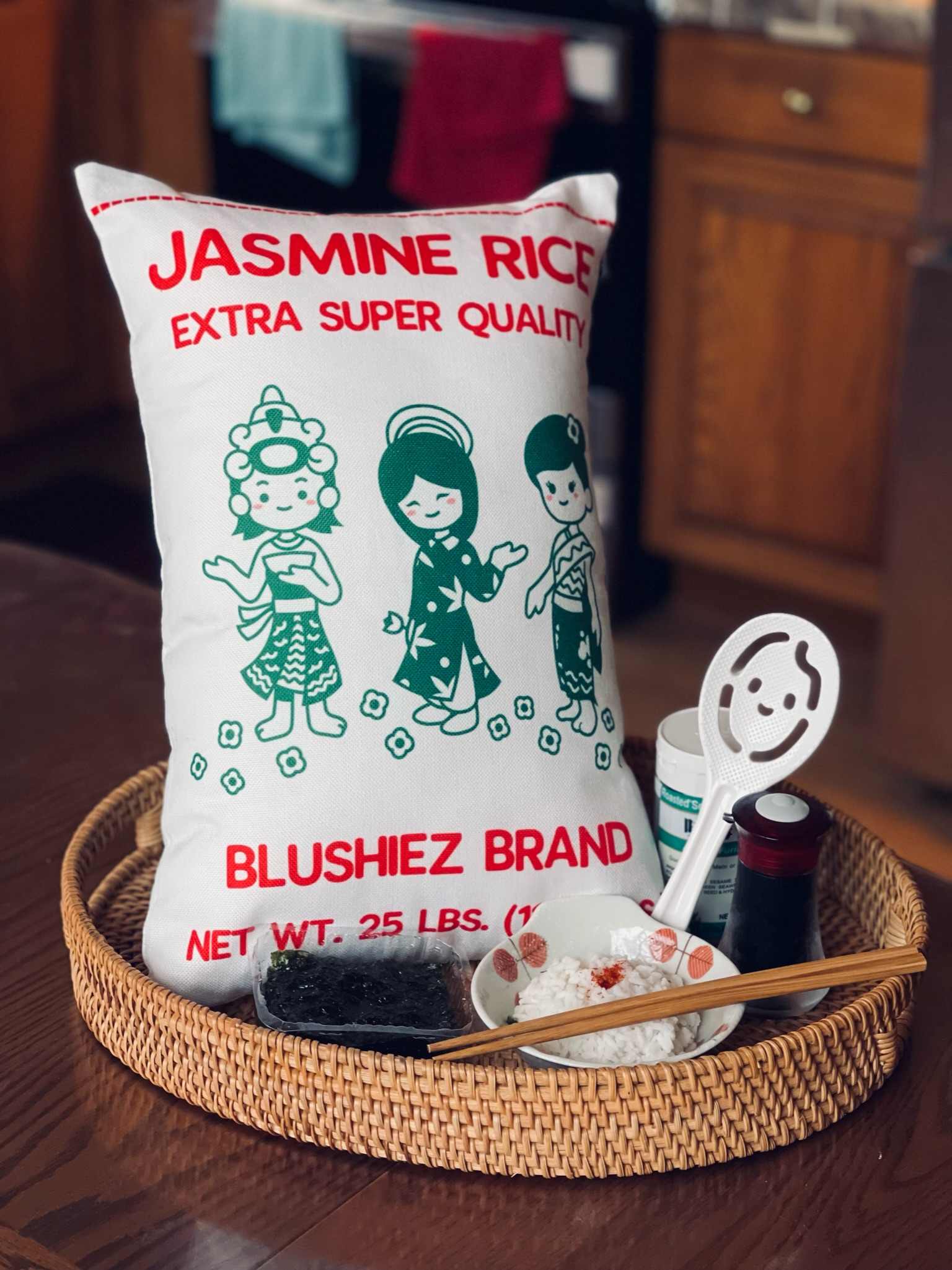Front of the pillow, resembling Jasmine Rice packaging, placed in a tray alongside a bowl of rice, soy sauce, and seaweed in a kitchen. The pillow features a kawaii-style illustration of three girls in green dresses on a white background, reminiscent of the vibrant imagery found on Jasmine Rice packaging. The scene depicts a cozy kitchen setting with elements representing a delightful meal.