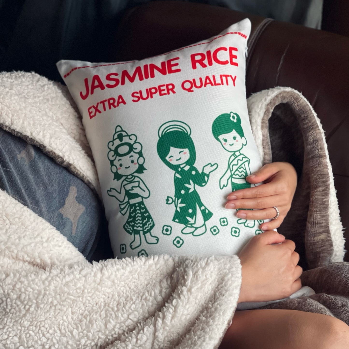 Front of the pillow, inspired by Jasmine Rice packaging, held in the hands of a woman in a seated position. The pillow features a kawaii-style illustration of three girls in green dresses on a white background. The playful design resembles the vibrant and culturally inspired imagery found on Jasmine Rice packaging. The woman's hands showcase the size and texture of the pillow, inviting a cozy and comforting experience.