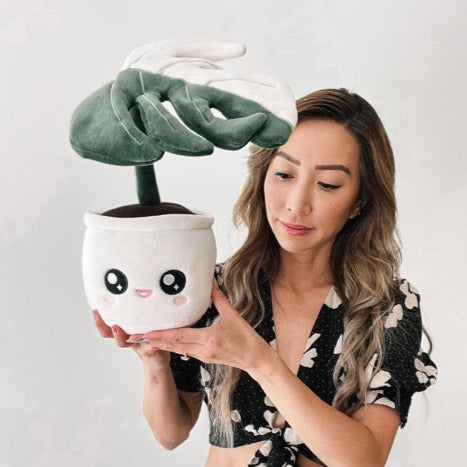 Plush toy of a potted plant with a white pot and a cute open-mouthed smiling face on the pot, being held with two hands by a woman. The plant is a single leaf of a Monstera deliciosa albo variegata, featuring a split color pattern. The left half of the leaf is green, while the right half is white.