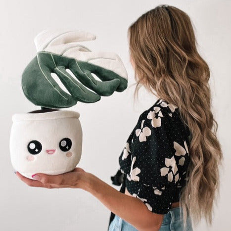Plush toy of a potted plant with a white pot and a cute open-mouthed smiling face on the pot, held by a woman's hand.