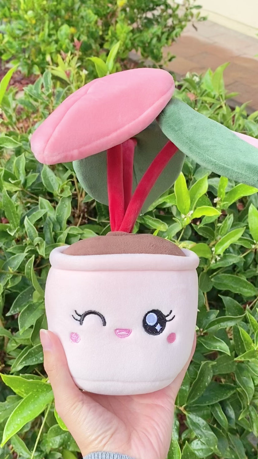 A video showcasing a woman holding a plush toy of a potted plant with a pink pot. The plant is a pink princess philodendron with three leaves. One leaf is green, another leaf is pink, and the third leaf has a split color pattern with the left side green and the right side pink. The plant's dark red stems are visible as the woman rotates the toy for the camera, providing different angles to appreciate the details of the plant.