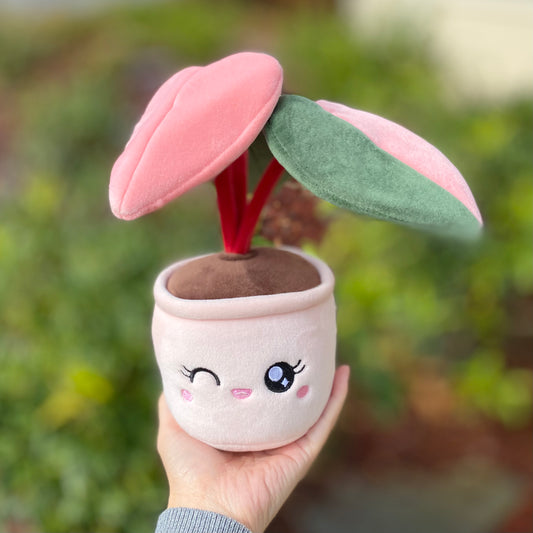 Plush toy of a potted plant with a pink pot and a winking face with eyelashes, held by a woman's hand in front of a bush. The plant is a pink princess philodendron with three leaves. One leaf is green, another leaf is pink, and the third leaf has a split color pattern with the left side green and the right side pink. The stems of the plant are dark red.