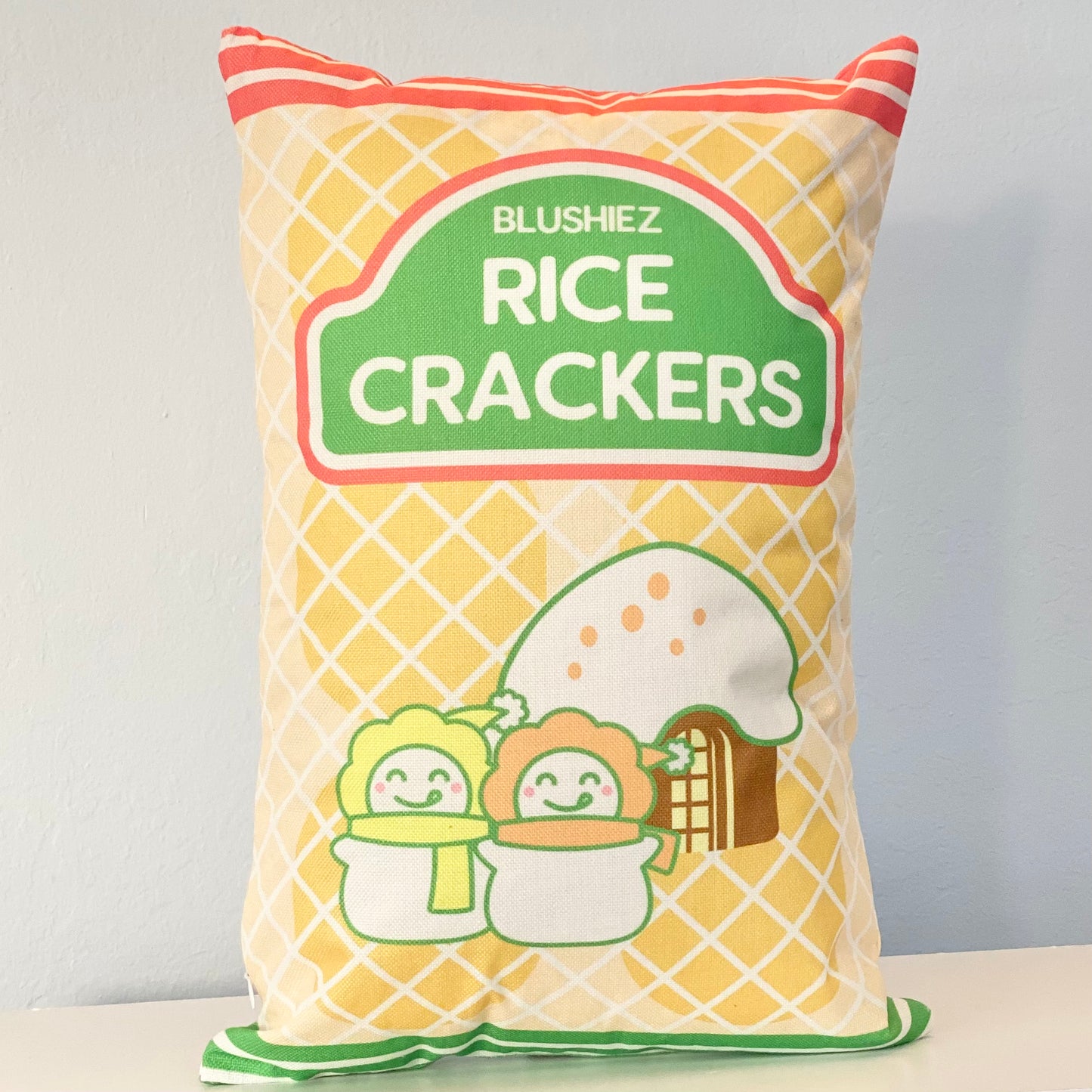 Pillow with a festive snack-inspired design. The pillow features a light orange background resembling rice crackers, with orange stripes at the top and green stripes at the bottom. The design includes a charming scene of two snowmen in front of a house with a snowy roof, evoking a festive and wintery atmosphere.