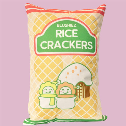 Pillow with a festive snack-inspired design. The pillow features a light orange background resembling rice crackers, with orange stripes at the top and green stripes at the bottom. The design includes a charming scene of two snowmen in front of a house with a snowy roof, evoking a festive and wintery atmosphere.