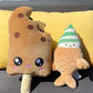 Plush toy of a boba ice cream bar with a smiling face, sitting on an outdoor sofa alongside a plush toy of a taiyaki soft serve ice cream with green and white swirls.