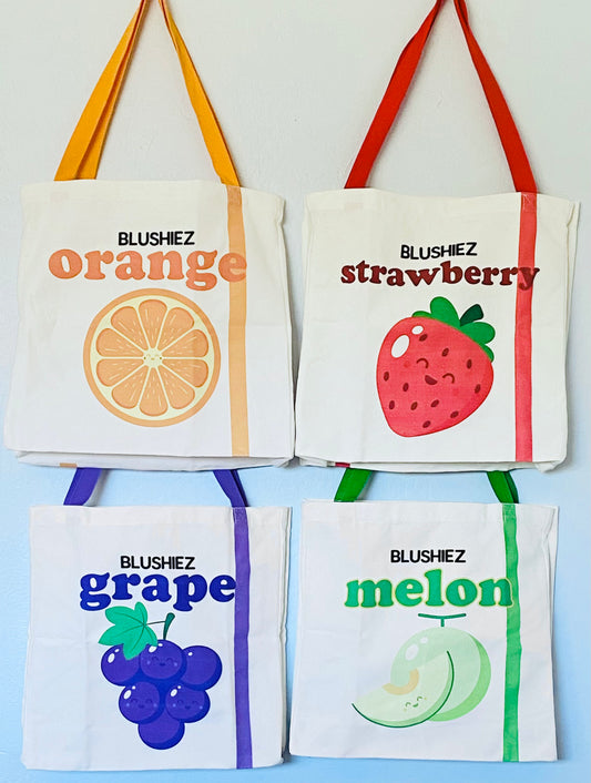 Four fruit themed tote bags each featuring a different fruit with a cute smiling face. Clockwise from top left: orange fruit with orange handle, strawberry fruit with red handle, grape fruit with purple handle, melon fruit with green handle.