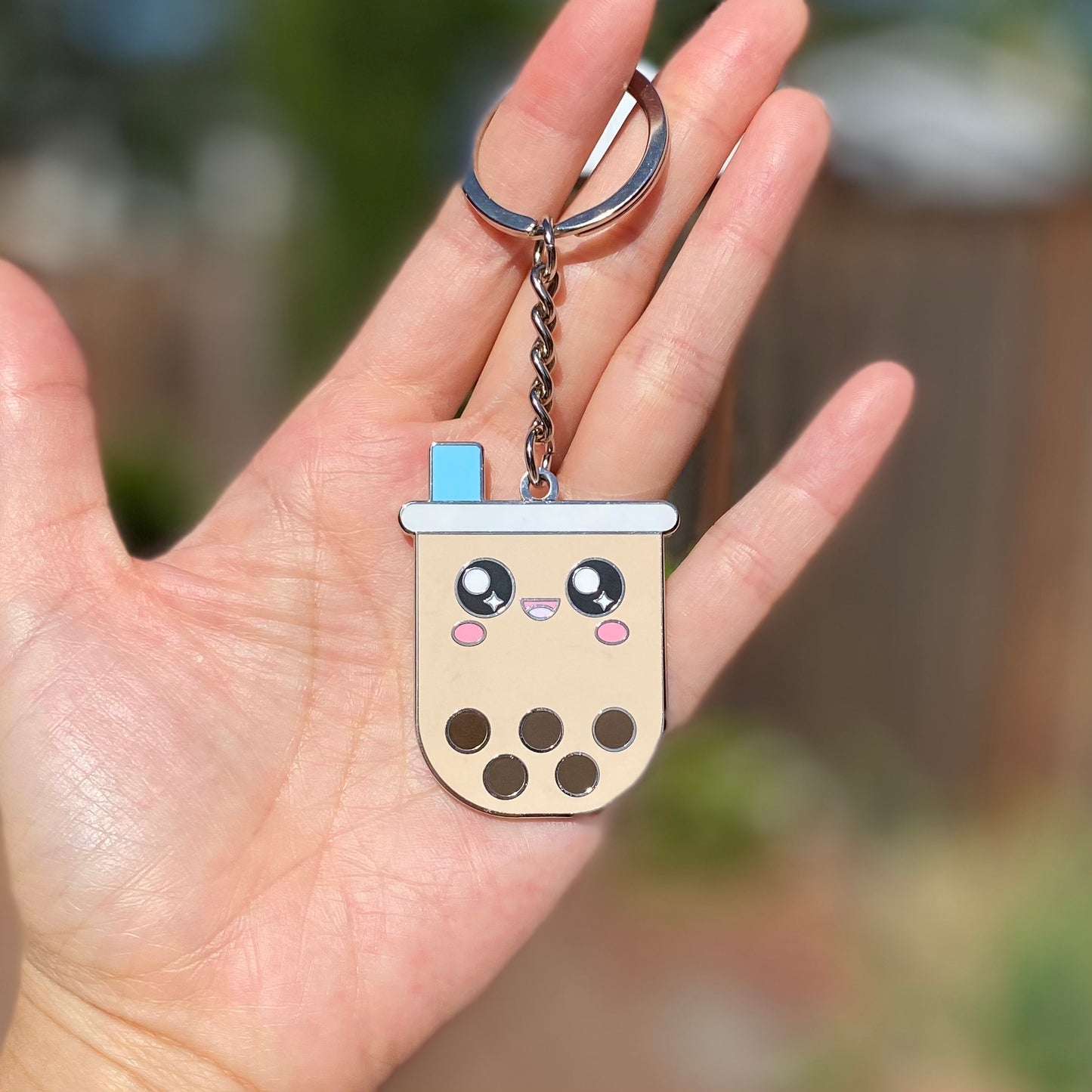 Enamel keychain held by a woman's hand. The keychain features a boba drink character with a cheerful face, a tapioca pearl-filled drink cup, and a blue wide straw.