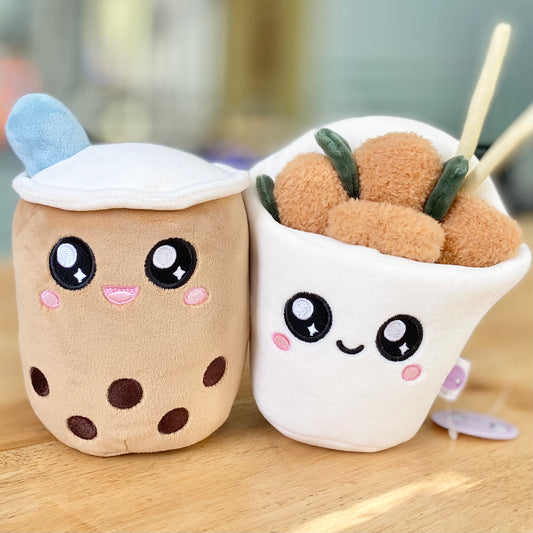 Plush toys of a boba drink and popcorn chicken characters placed on a countertop. The boba drink character features a cute smiling face, while the popcorn chicken character also has a cheerful expression.