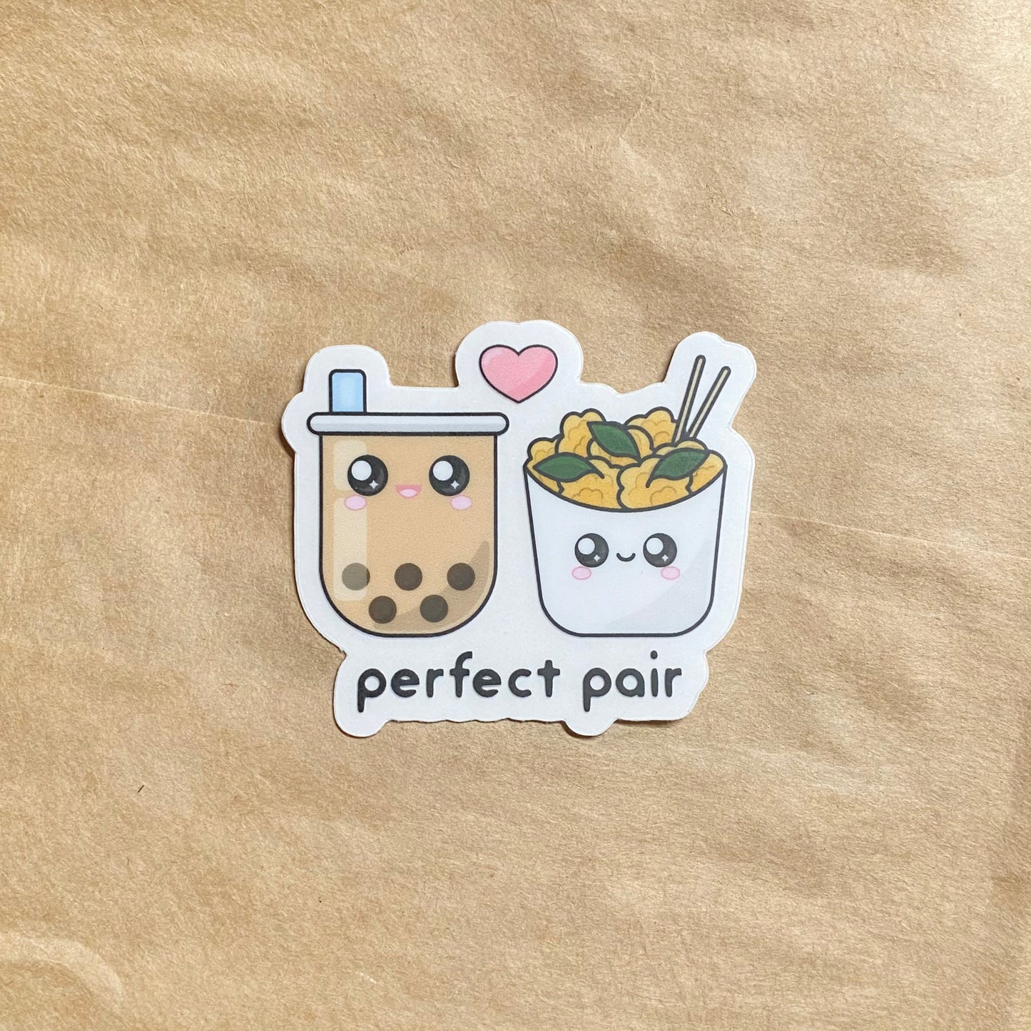 Sticker with a clear background placed on a brown piece of paper, showing the white paper backing still attached. The sticker features a cute open-mouth smiling boba milk tea and a closed-mouthed smiling Taiwanese popcorn chicken. A pink heart is between them, with the words 'perfect pair' printed below.