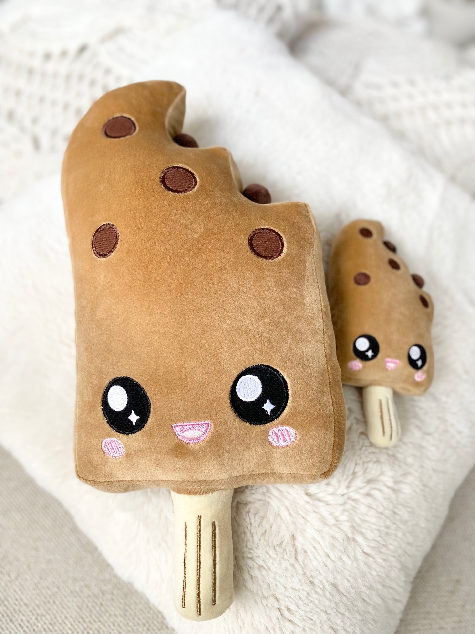 Plush toy of a boba ice cream bar with a cute open-mouth smiling face lying next to a miniature version of itself on a fluffy white pillow.