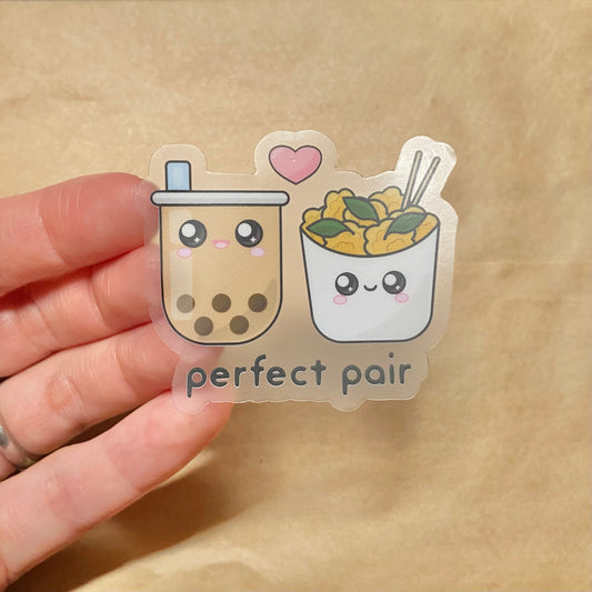 Sticker with a clear background partially stuck to the fingers of a woman's hand. The sticker features a cute open-mouth smiling boba milk tea and a closed-mouthed smiling Taiwanese popcorn chicken. A pink heart is between them, with the words 'perfect pair' printed below.