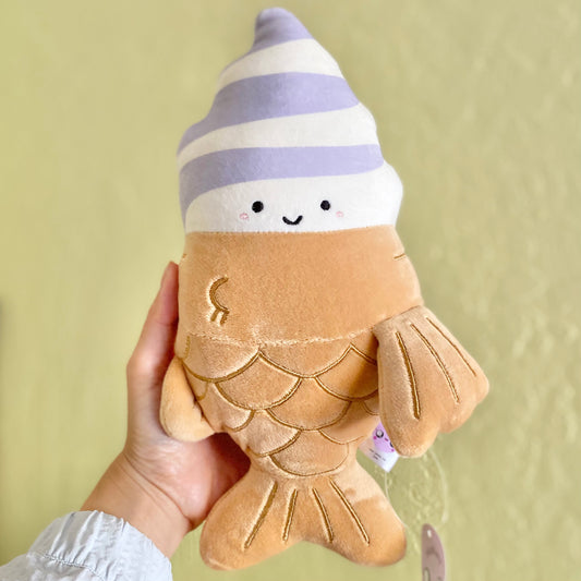 Plush toy of a Taiyaki ice cream with a fish-shaped cone body and a soft serve swirl head featuring purple and white swirls, held by a woman's hand.