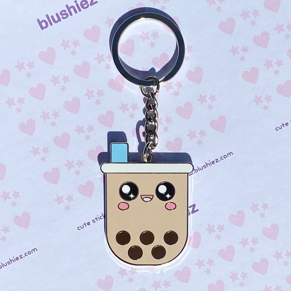 Enamel keychain laying on a white background with a pattern featuring pink hearts and stars. The keychain features a boba drink character with a cheerful face, a tapioca pearl-filled drink cup, and a wide straw.