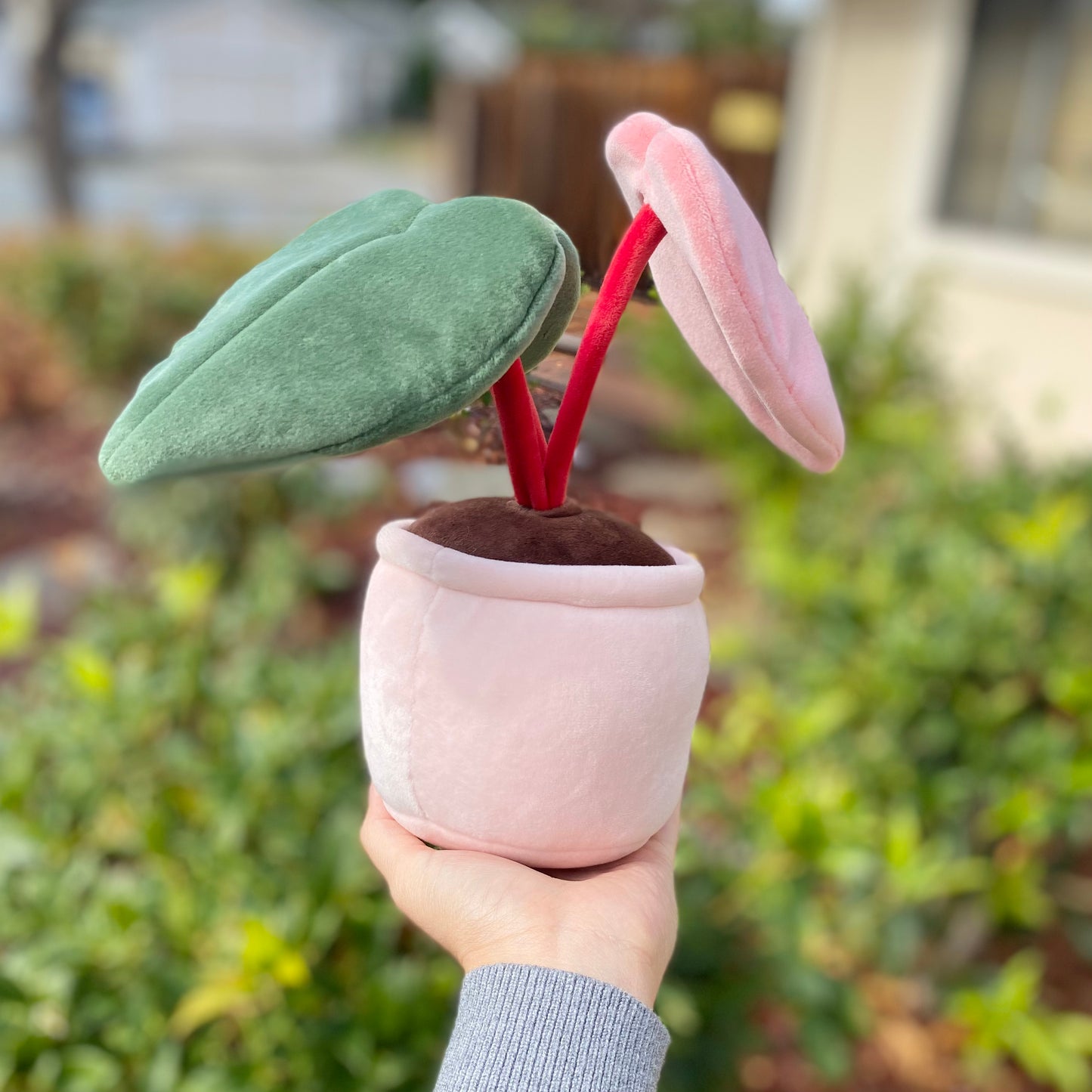 Rear view of the plush toy of a potted plant with a pink pot, held by a woman's hand in front of a bush. The plant is a pink princess philodendron with three leaves. One leaf is green, another leaf is pink, and the third leaf has a split color pattern with the left side green and the right side pink. The dark red stems of the plant are visible.
