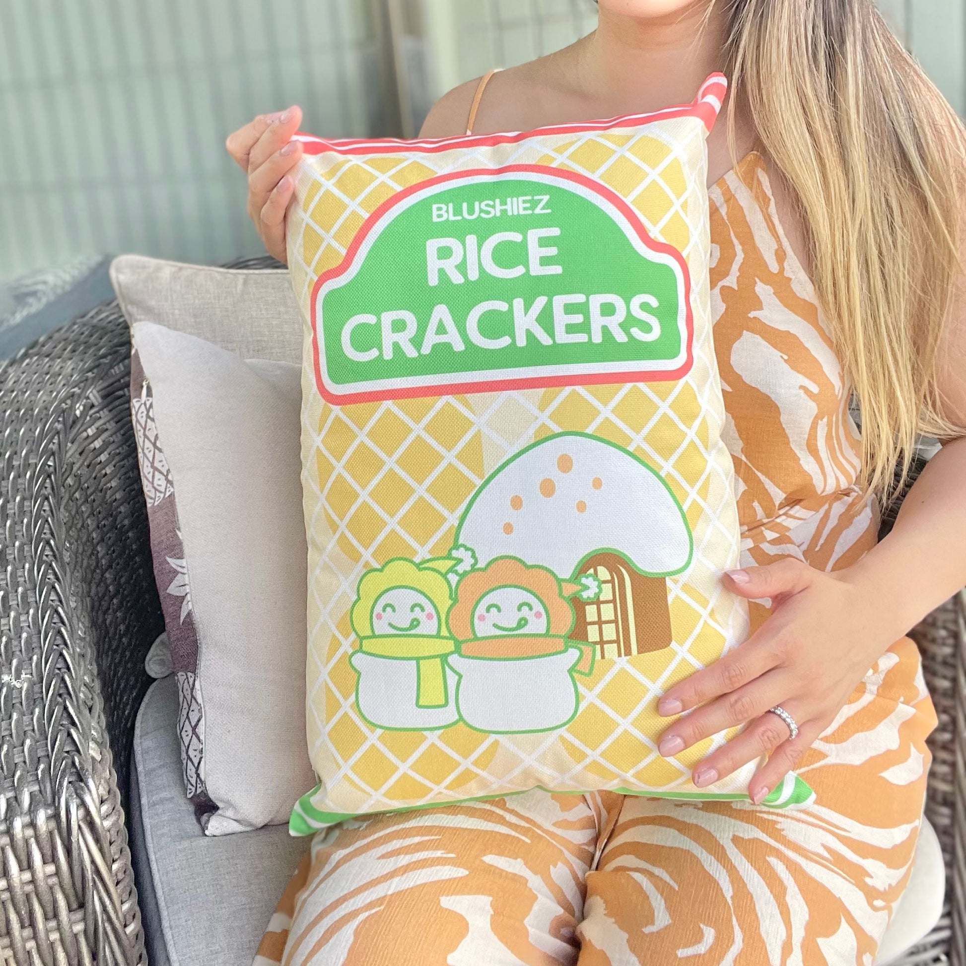 Pillow with a festive snack-inspired design, held by a woman in her lap. The pillow features a light orange background resembling rice crackers, with orange stripes at the top and green stripes at the bottom. The design includes a charming scene of two snowmen in front of a house with a snowy roof, evoking a festive and wintery atmosphere.