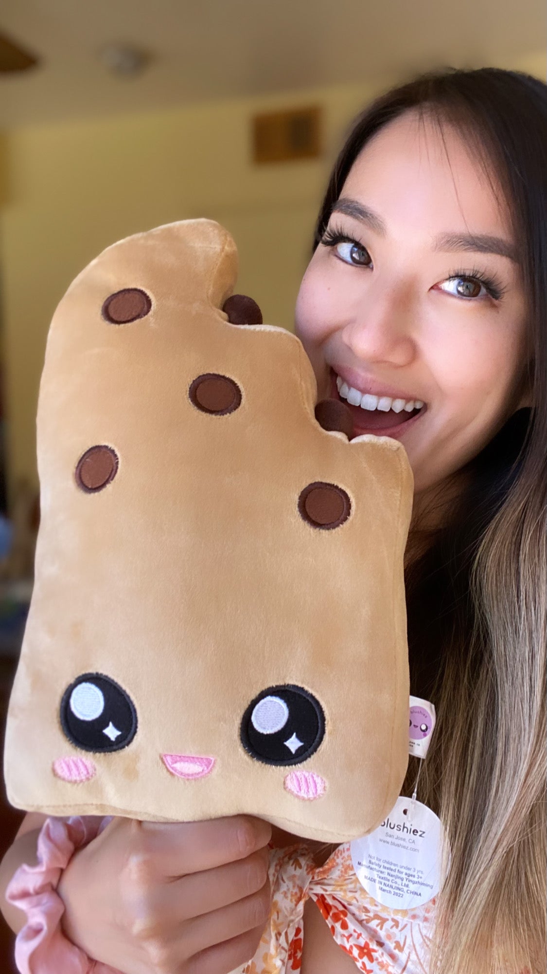 Woman holding a plush toy of a boba ice cream bar with a cute open-mouth smiling face. The plush toy is shaped like an ice cream bar with a bite taken out of the top right corner. The woman is smiling and appears to be taking a playful bite out of the corner.